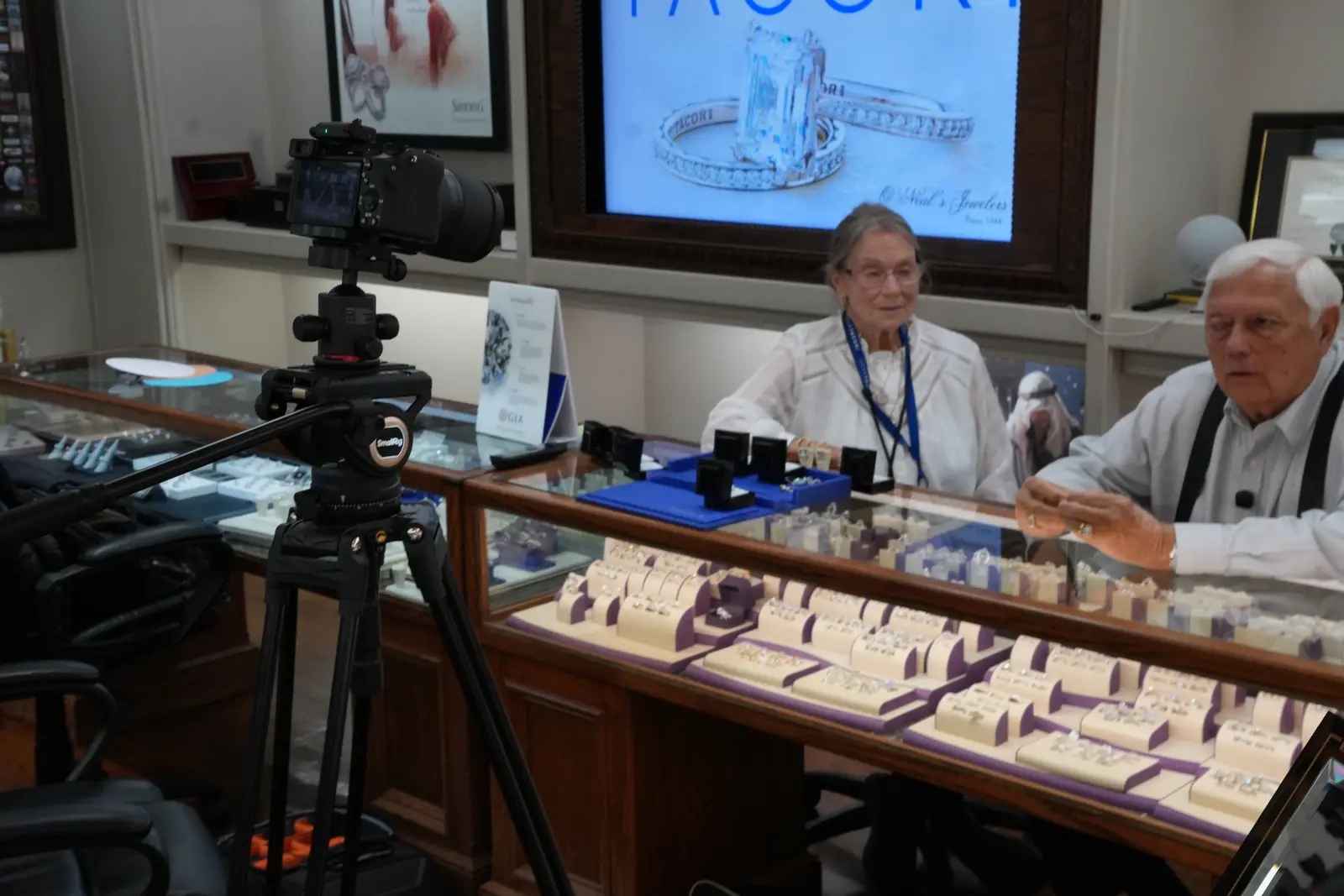 A camera on a tripod filming the owners of O'Neal's Jewelers in Pauls Valley, Oklahoma, at their counter, with a Tacori display behind them