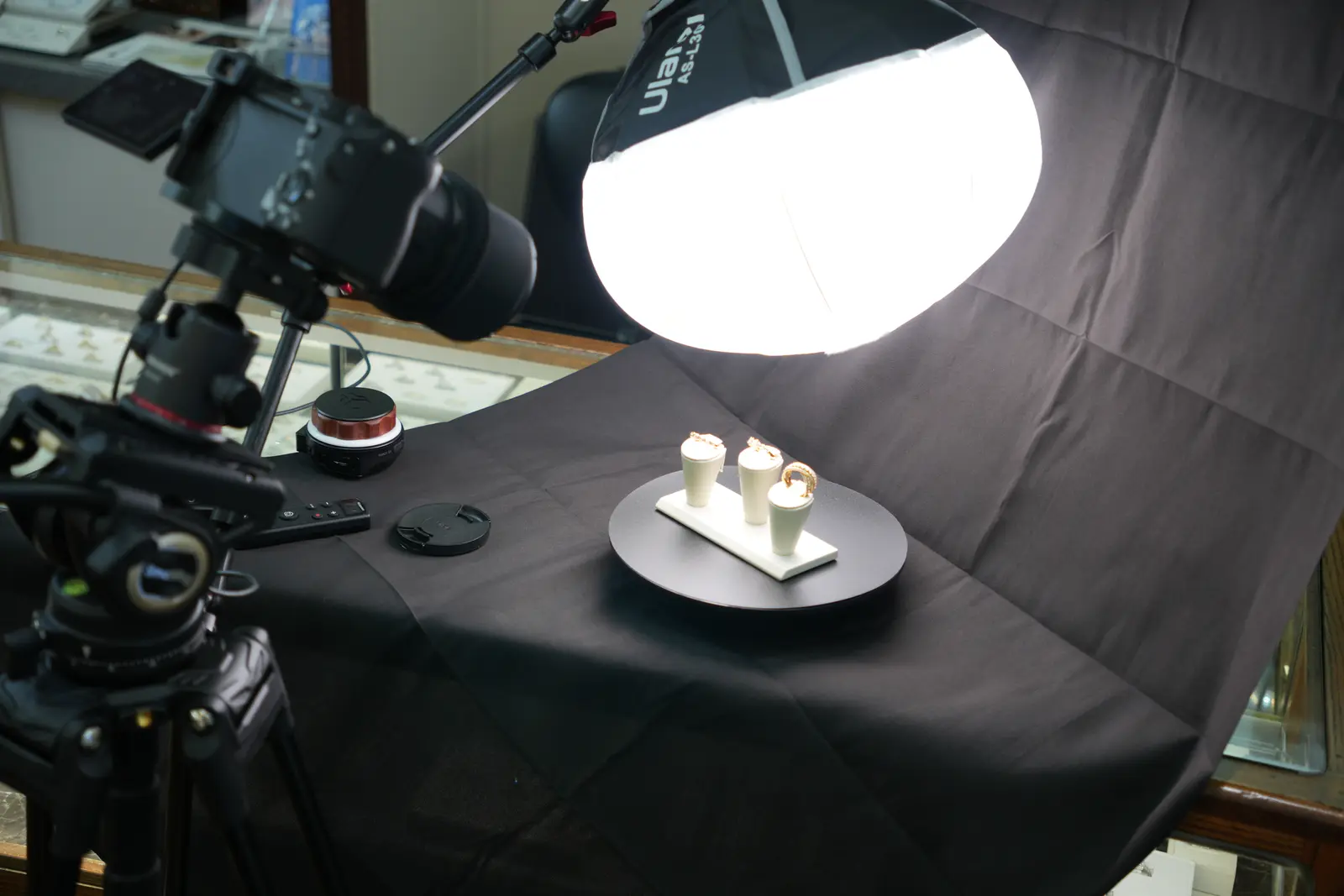 Ring stands on a black turntable under a large round softbox, with a camera on a tripod beside them