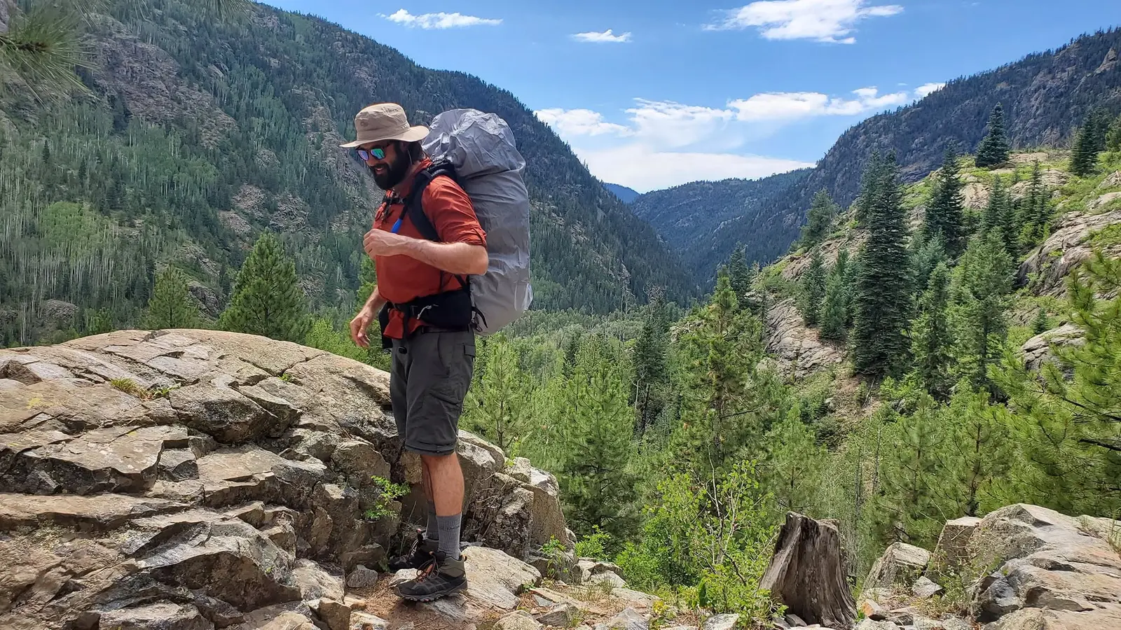 Mitch with a full backpack standing on a rocky ledge above a forested mountain valley