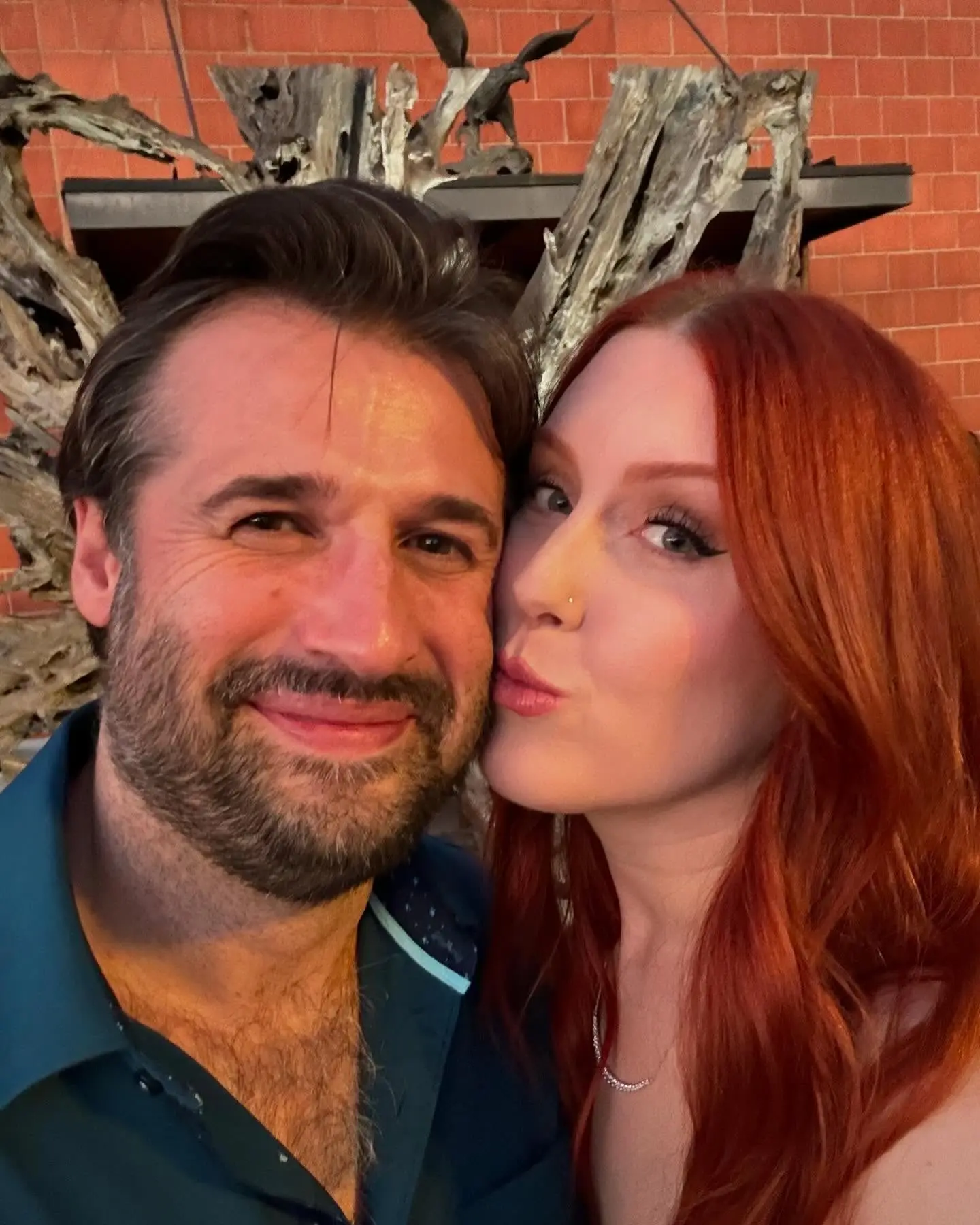 Close selfie of Mitch and Brittany Walker against a brick wall, Brittany kissing his cheek