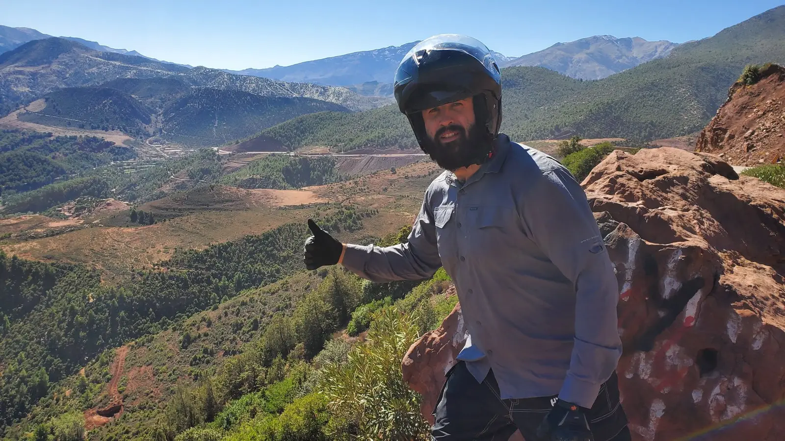 Mitch in a motorcycle helmet giving a thumbs-up on a trail above a canyon