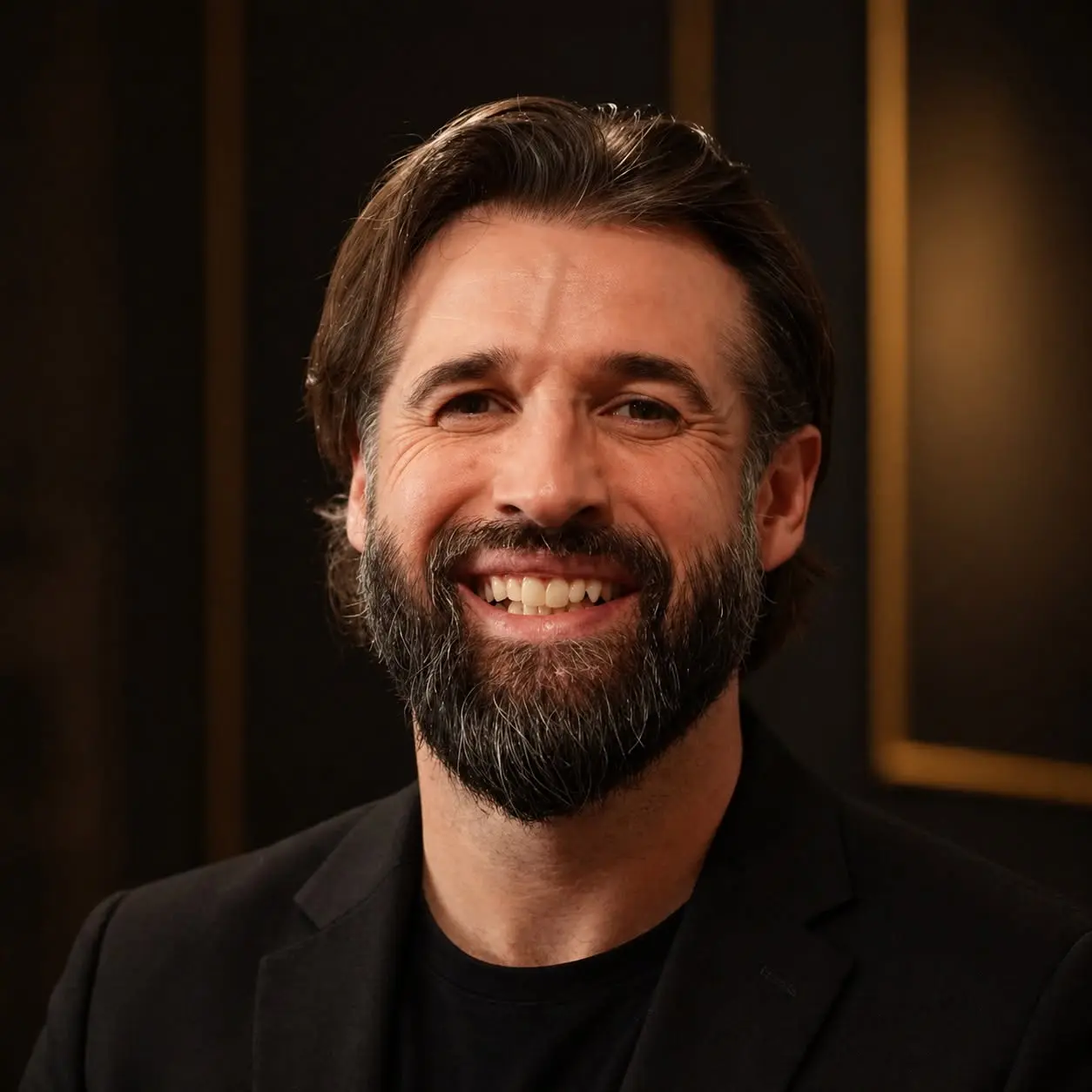 Mitch Walker smiling, with a full beard, in a black blazer and black T-shirt against a dark background with gold trim